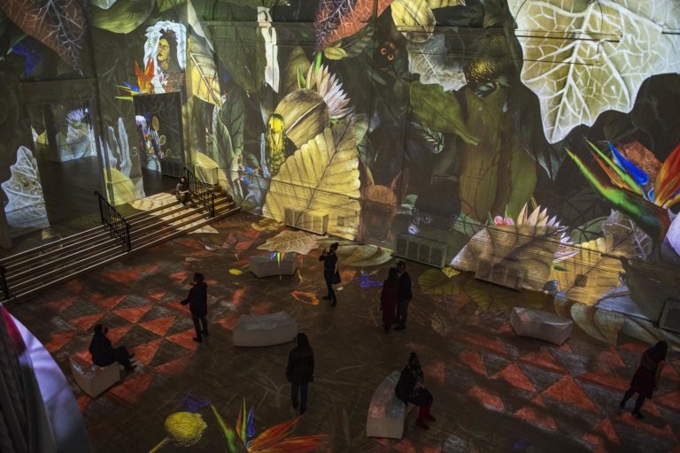 Photo from the Immersive Frida Kahlo exhibition showing Kahlo's artwork projected on the wall and spectator silhouettes in the foreground
