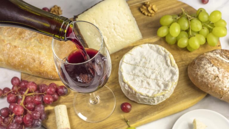 Red wine poured into a glass at a tasting, with various types of cheeses, bread, and grapes.
