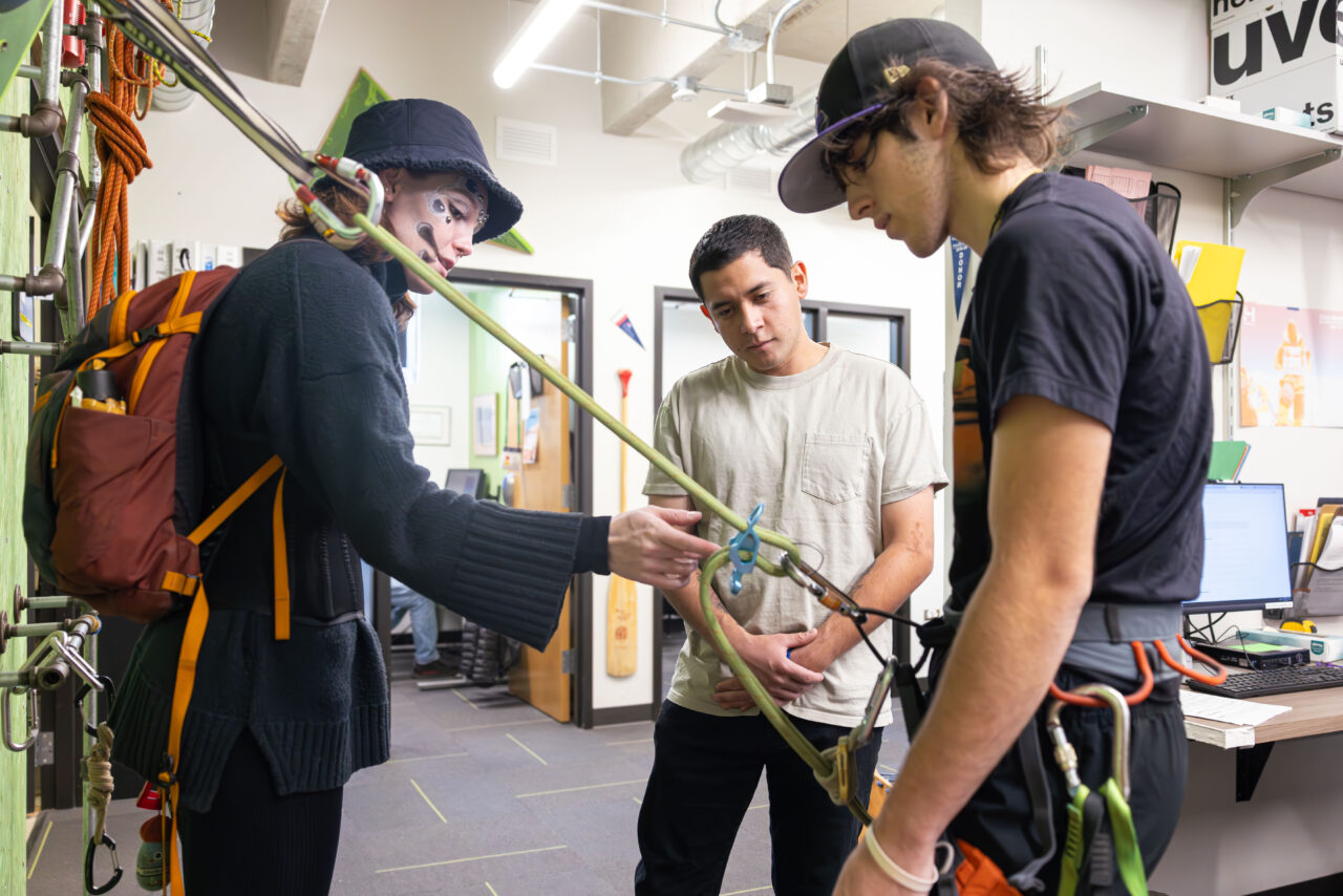 Three individuals discussing climbing equipment setup in the Outdoor Pursuits office.