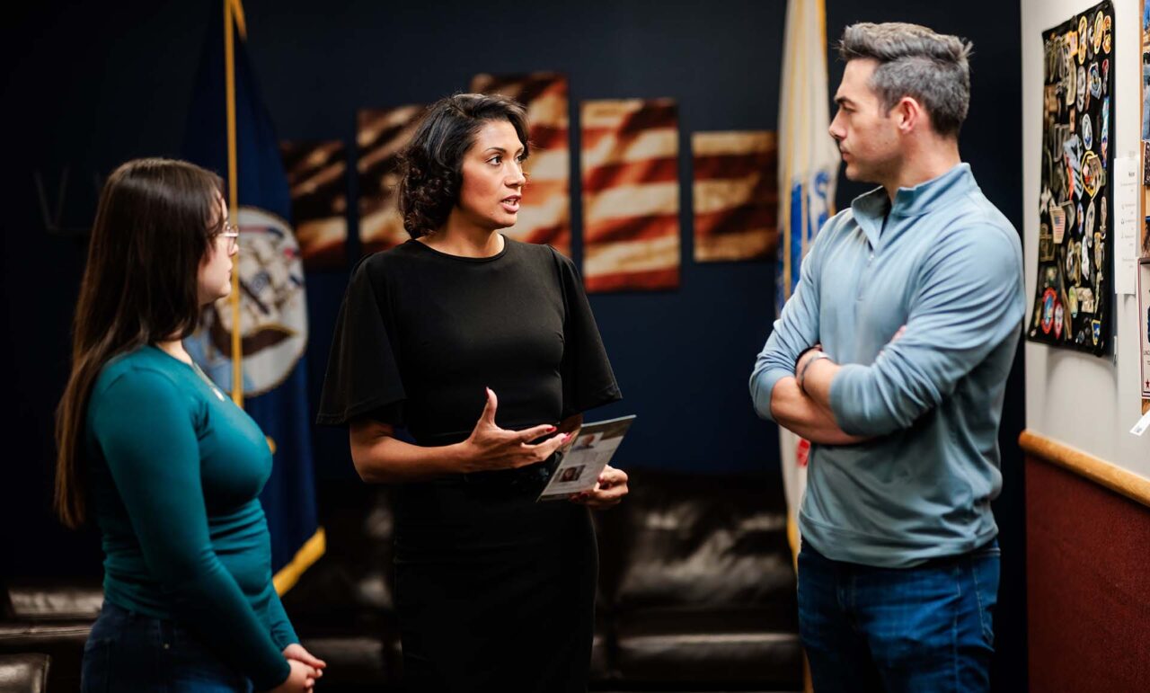 Leah Jackson, in a black dress, speaks with veterans Evelyn Beltran and Garrett Harlan in an office adorned with American flags and military memorabilia.