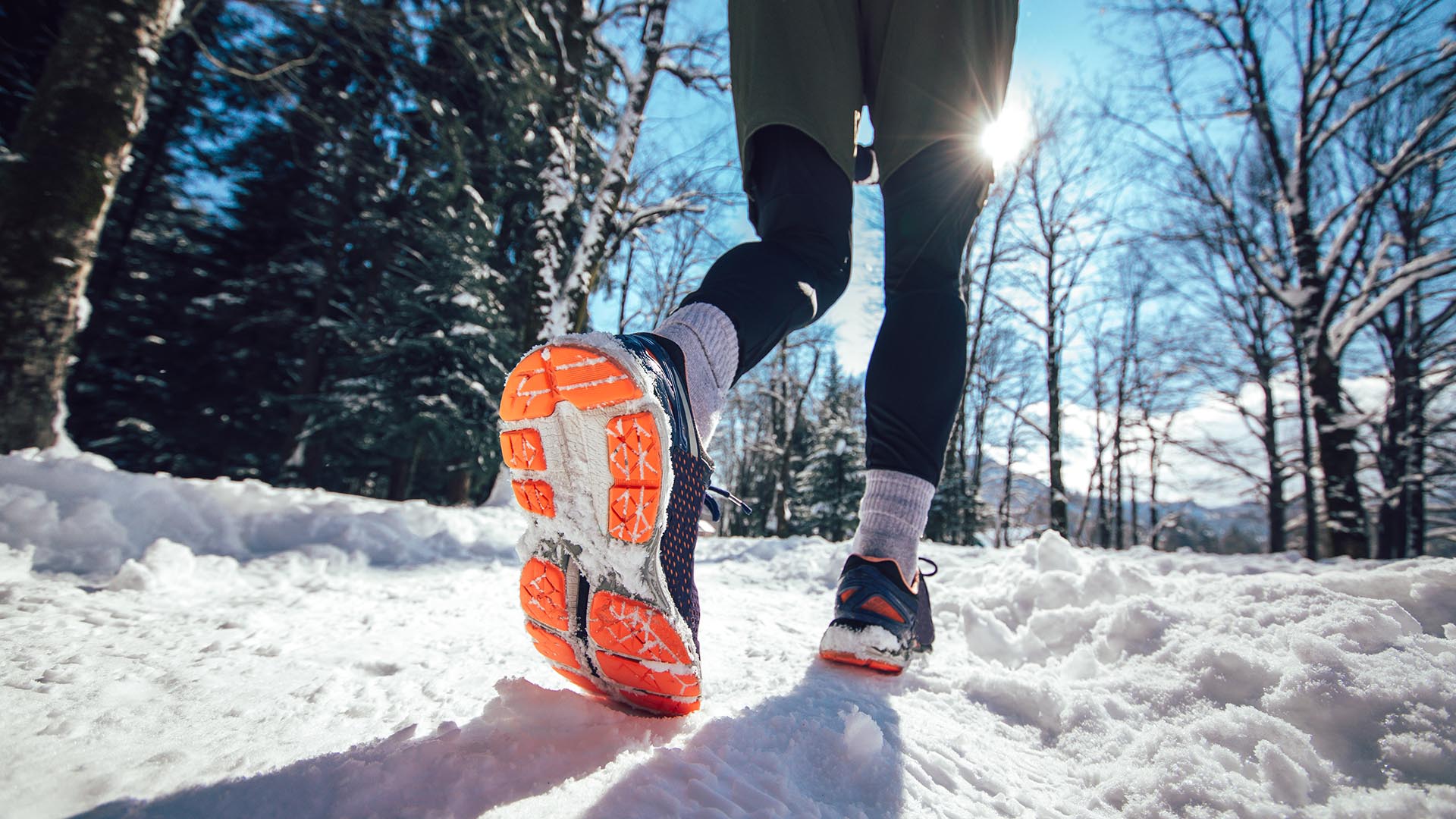 ALT Text: A runner in bright orange and black shoes jogs through a snow-covered forest trail under a clear blue sky.