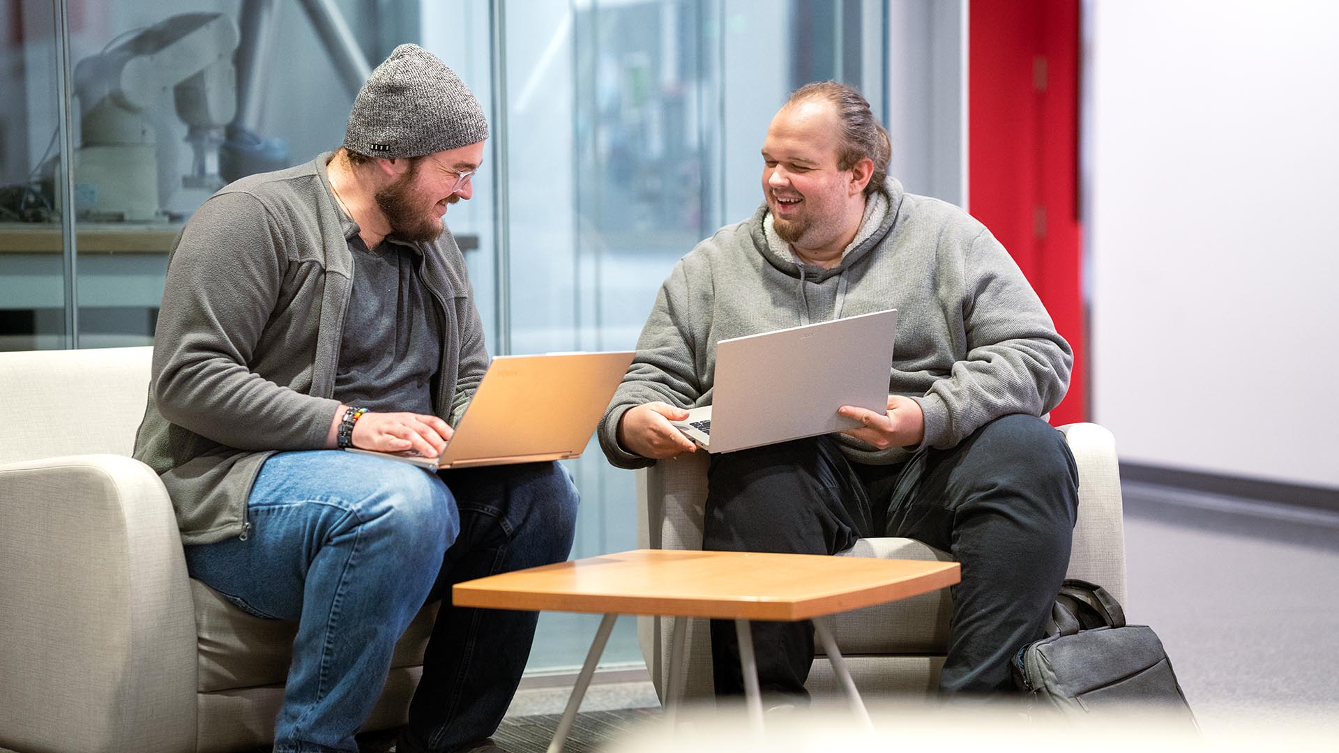 Two young men sit on a couch with laptops, smiling and talking in a modern lounge. A glass wall behind them reveals a workspace with a robotic arm.