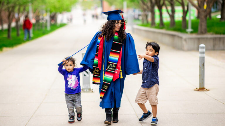 A woman wearing a blue graduation cap and gown walks down a tree-lined campus sidewalk holding hands with two young children. She wears colorful stoles, including one embroidered with “Jaclyn Gutierrez” and “MSU Denver.” A little girl in a purple unicorn shirt walks on her left, playfully holding a graduation tassel string, while a young boy in a navy blue polka-dotted shirt and khaki shorts walks on her right, pointing ahead excitedly.