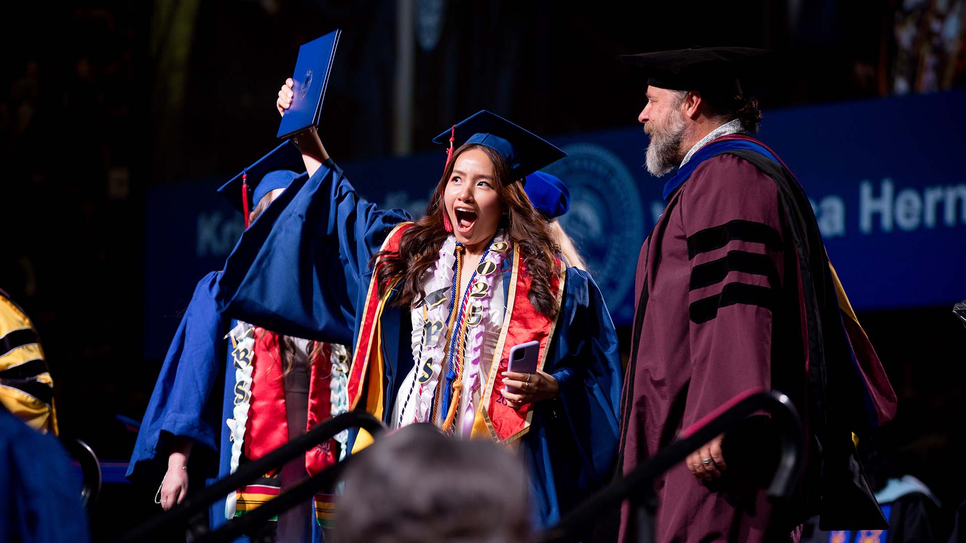 Smiling graduate raises her diploma as she walks across the stage