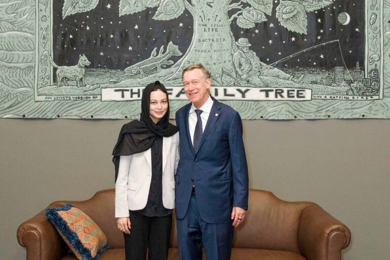 Madina Amiri stands next to Senator John Hickenlooper, both smiling in front of a banner labeled “The Family Tree.”