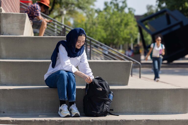 A student wearing a navy hijab and white shirt sits on outdoor steps, smiling while opening a black backpack.