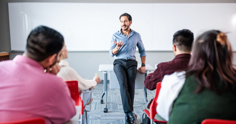 An instructor sits on a desk, speaking to students in a classroom.