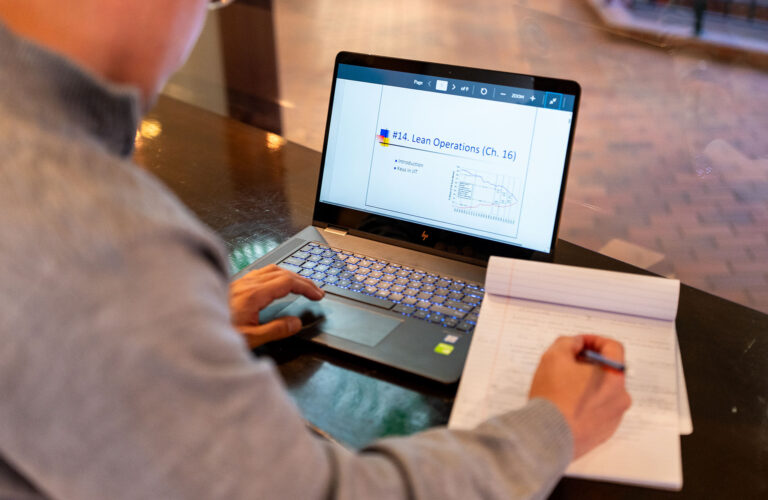 A person takes notes while viewing a “Lean Operations” lecture slide on a laptop.