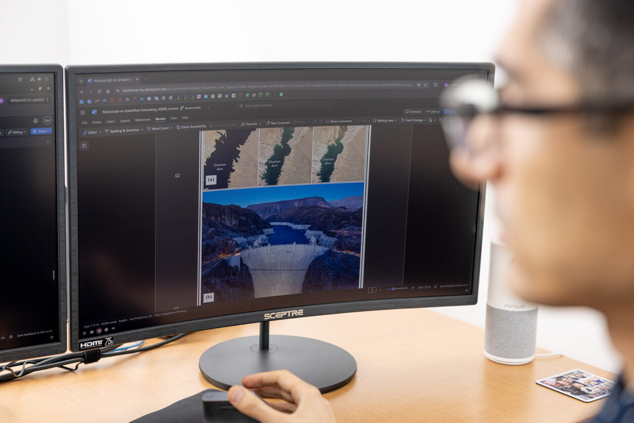 A person looks at a computer monitor displaying maps and a photo of a dam while taking notes with a mouse in hand.