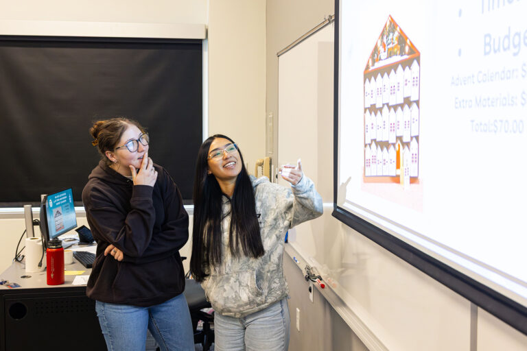 Two students stand at the front of a classroom, smiling and discussing a presentation projected on a screen.