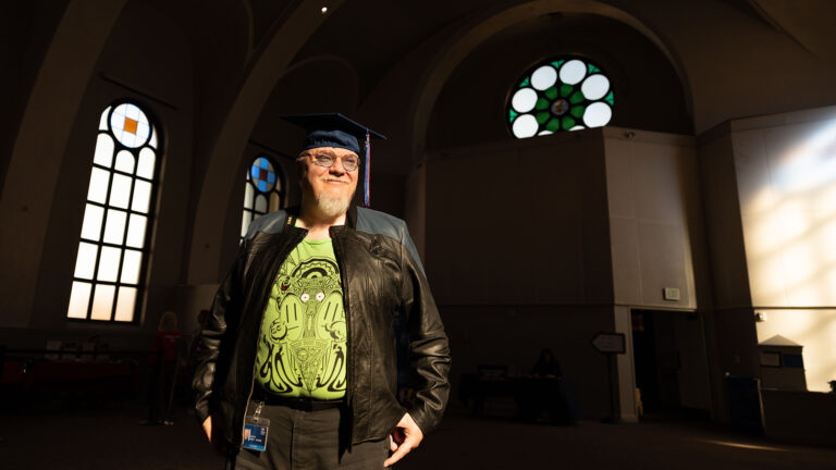 A person wearing a graduation cap stands in a shaft of light inside St. Cajetan’s, smiling confidently.