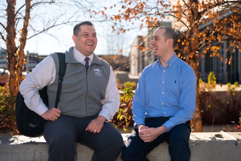 Two people sit outdoors on a concrete ledge, smiling and talking on a sunny day.