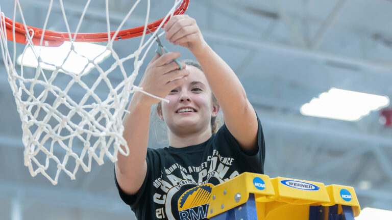 A student athlete stands on a ladder cutting down a basketball net after a championship win.
